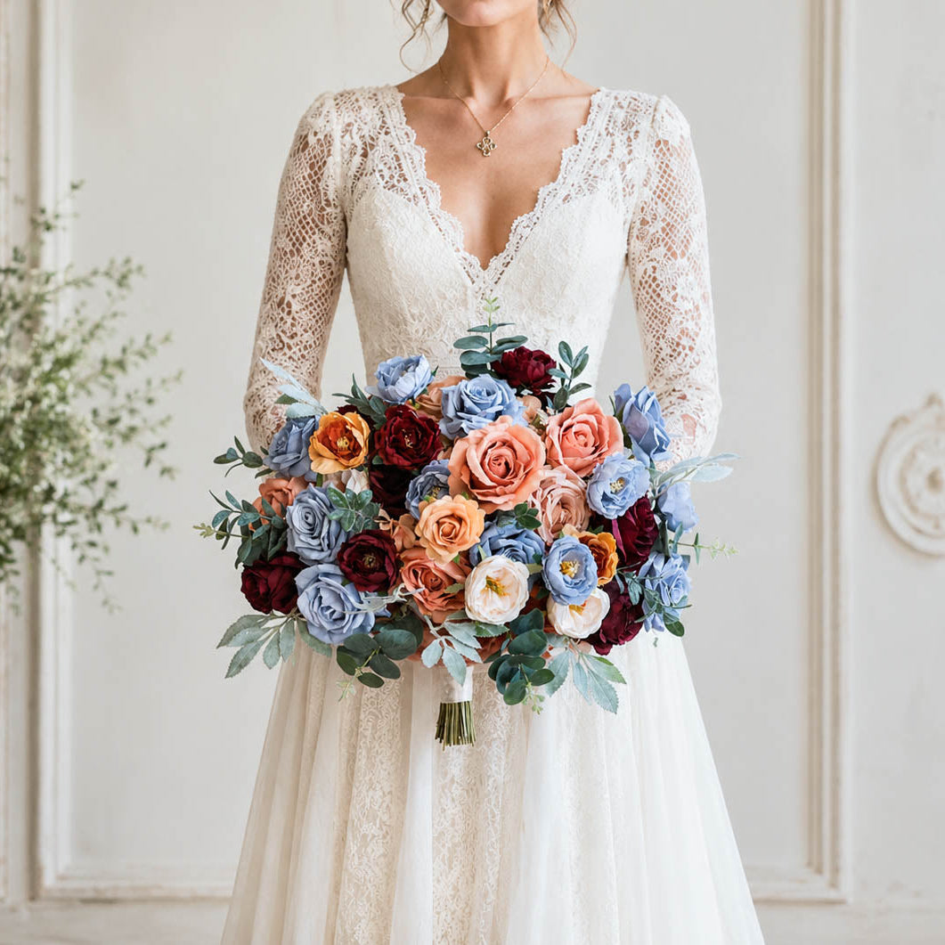 A bride in a white lace wedding dress holding a colorful bouquet of blue, orange, burgundy, and cream flowers, standing in a classic white interior with decorative wall molding.