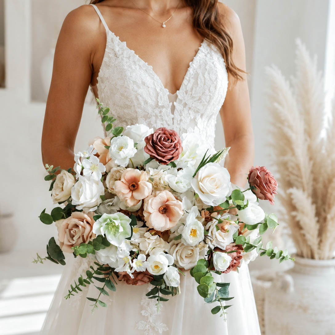 A bride in a white lace wedding dress holding a large bridal bouquet with cream, blush, and dusty rose flowers and green foliage, photographed in a bright indoor setting with pampas grass in the background.