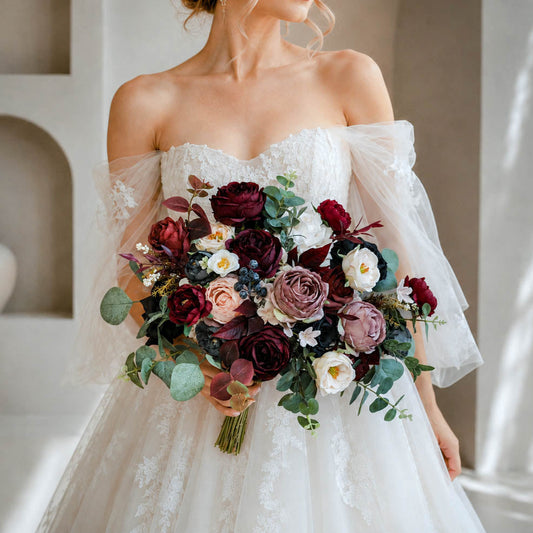 A bride wearing an off-the-shoulder white lace wedding dress holds a large bridal bouquet of burgundy, mauve, white, and green flowers, standing in a softly lit interior with architectural recesses and window shadows.