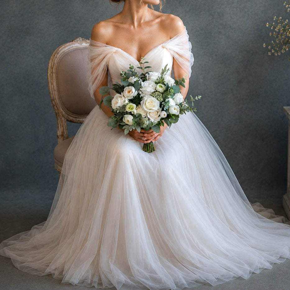 A woman seated on a vintage upholstered chair wearing an off-the-shoulder white wedding gown with a full tulle skirt and holding a bouquet of white flowers and green leaves, posed against a dark gray studio backdrop with a stone side table and floral arrangement beside her.